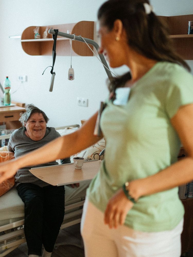 A carer greeting a smiling older woman sitting on her bed
